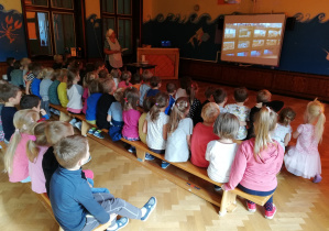 Dzieci ze starszych wraz z prowadząca spotkanie oglądają film edukacyjny na tablicy multimedialnej w sali gimnastycznej